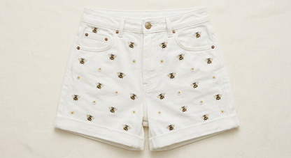 Detailed close-up showing the artisanal honeybee and daisy embroidery on white denim summer shorts.