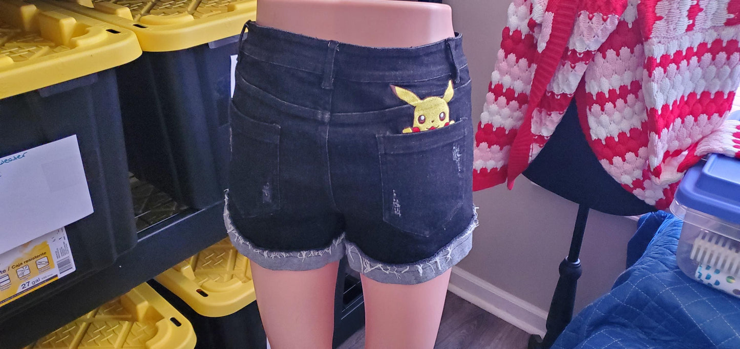 High-waisted black cotton denim shorts featuring Pikachu anime embroidery on the back pocket and a raw frayed hem.