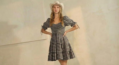 Woman in a black gingham puff sleeve mini dress with a shirred bodice and tiered skirt, wearing a western cowboy hat.