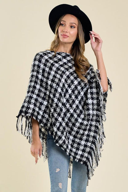 Women's black and white houndstooth print knit poncho featuring an asymmetric drape and fringe trim, styled with a black hat.