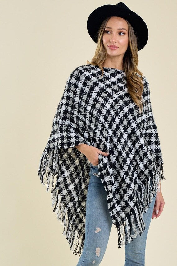 Woman wearing a black and white houndstooth knit poncho with fringe trim, styled with a black hat and blue jeans.