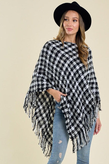 Woman wearing a black and white houndstooth knit poncho with fringe trim, styled with a black hat and blue jeans.