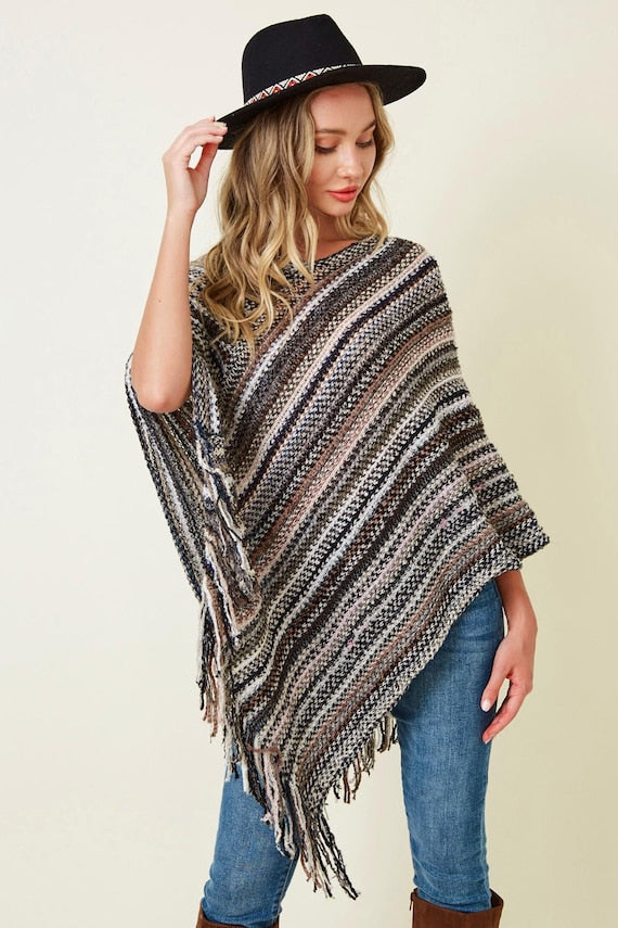 Woman wearing a striped knit poncho with fringe in neutral earth tones, styled with blue jeans and a black wide-brim hat.