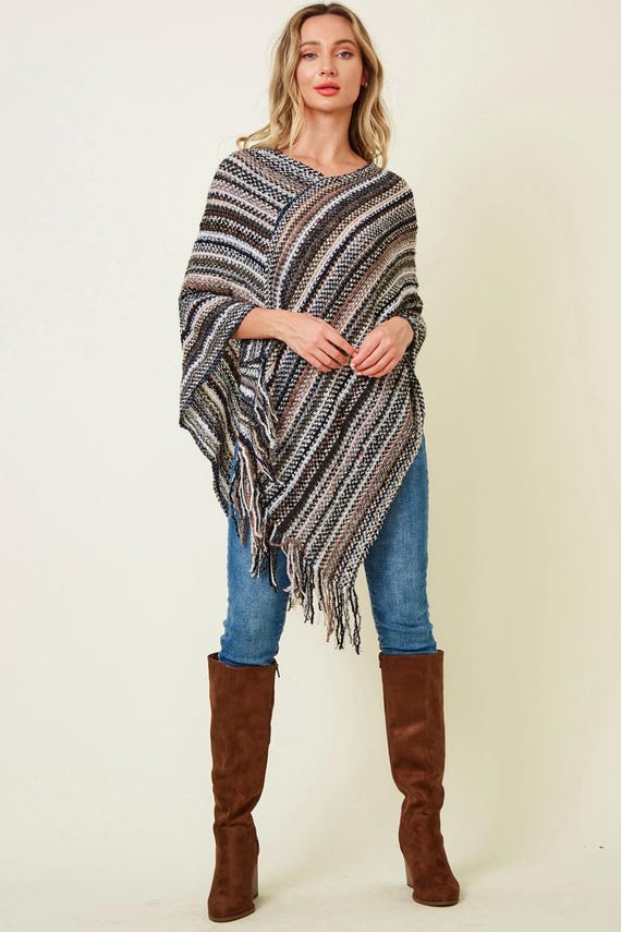 Woman wearing a striped knit poncho with fringe in neutral earth tones, paired with blue jeans and brown suede boots.