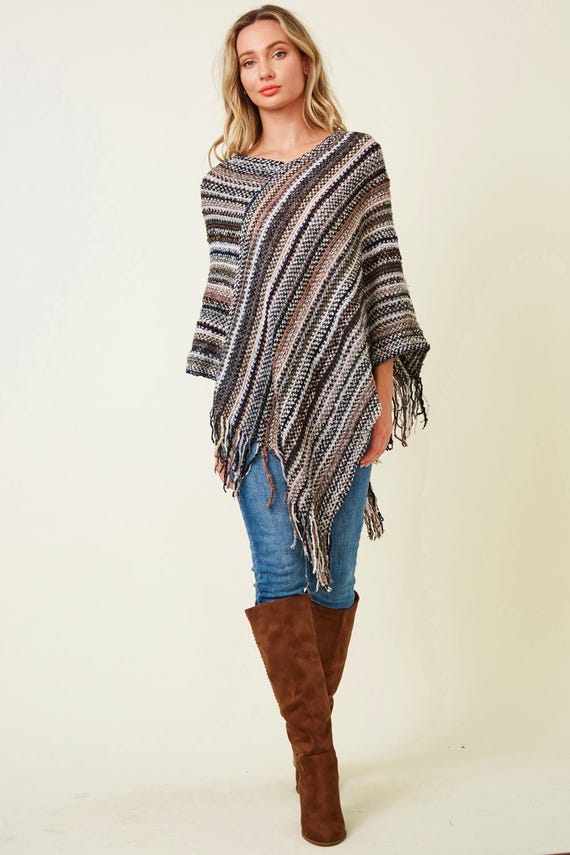 Women's striped knit poncho with boho fringe in neutral earth tones, paired with blue jeans and tall brown boots.