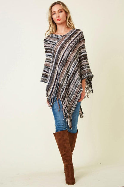 Women's striped knit poncho with boho fringe in neutral earth tones, paired with blue jeans and tall brown boots.