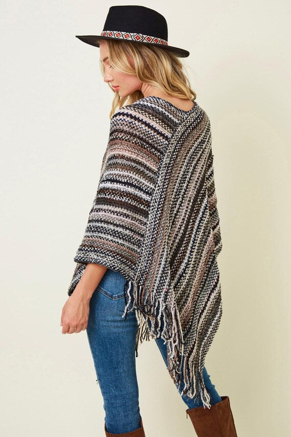 Woman wearing a striped knit poncho with fringe in boho casual earth tones, styled with blue jeans and a black felt hat.