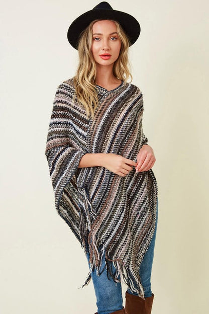 Women's boho casual striped knit poncho with fringe details in neutral earth tones, styled with blue jeans and a black hat.