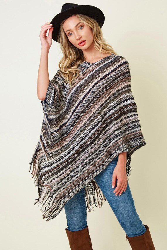 Women's boho striped fringe acrylic poncho in brown and grey knit, styled with blue jeans and a black wide-brim hat.