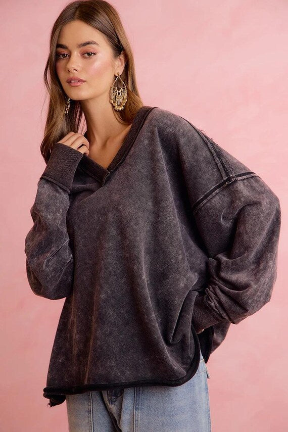 Model wears a mineral wash charcoal oversized sweatshirt with raw seams and dropped shoulders, v-neckline, and ribbed cuffs.